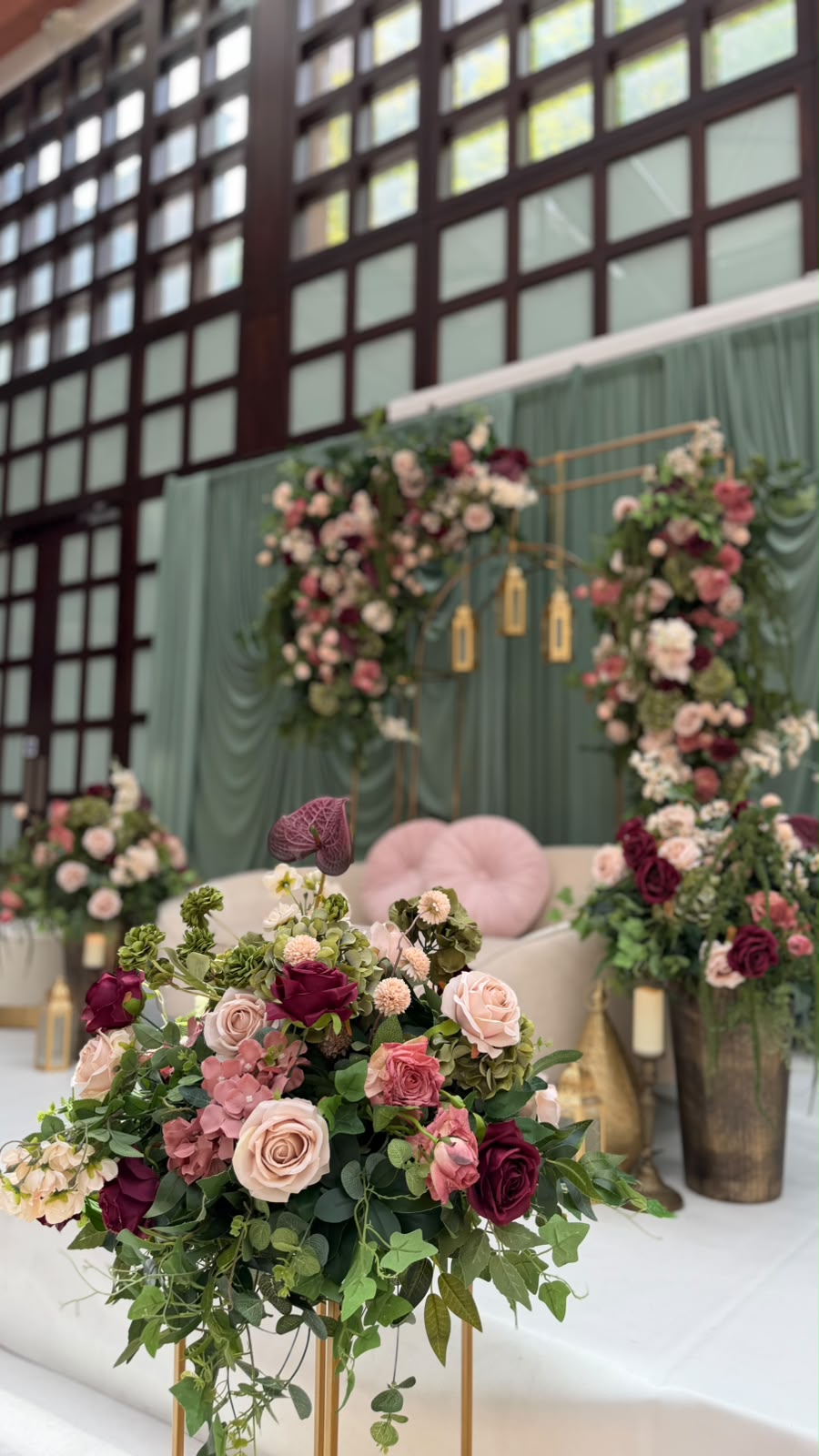 A stage against a sage green drape with two brass arches covered in roses, a blush sofa and tall floral stands